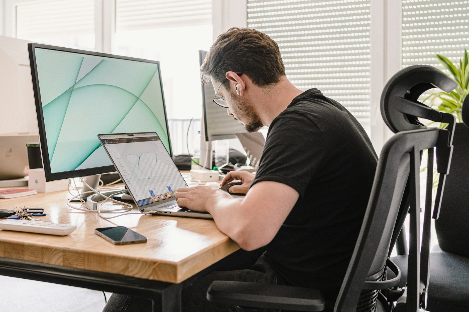 A man in a black T-shirt working on a laptop at a wooden desk beside a large monitor in a bright office