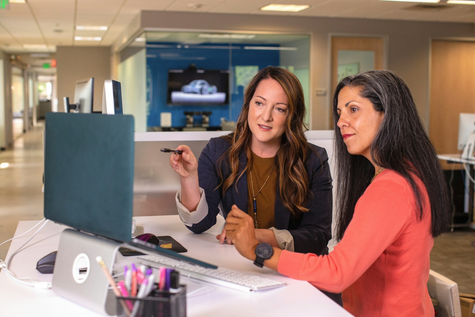 Two colleagues at an office desk look at a monitor together while one points at the screen with a pen