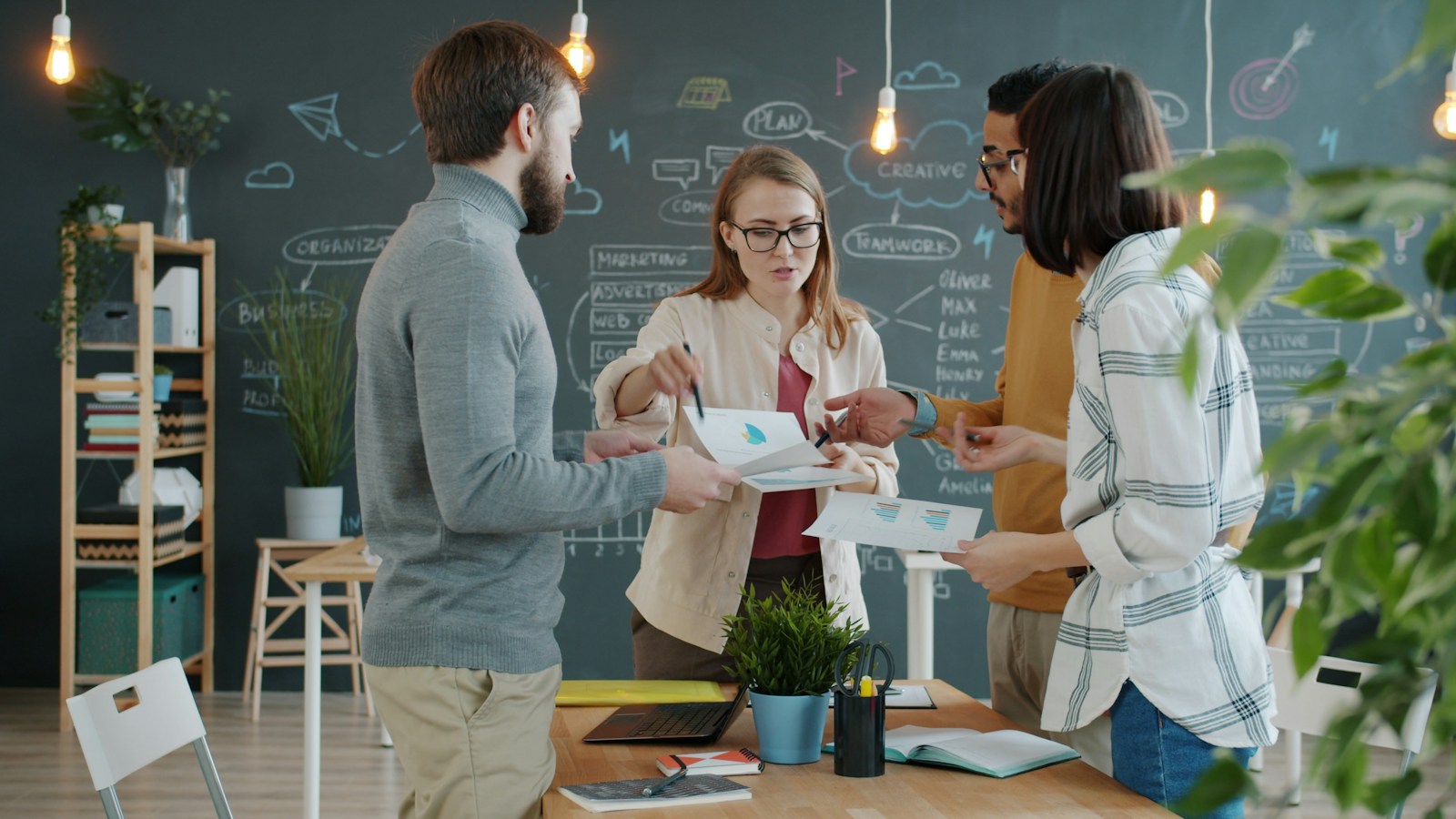 Four colleagues standing around a desk comparing printed charts, with a chalkboard of planning diagrams behind them