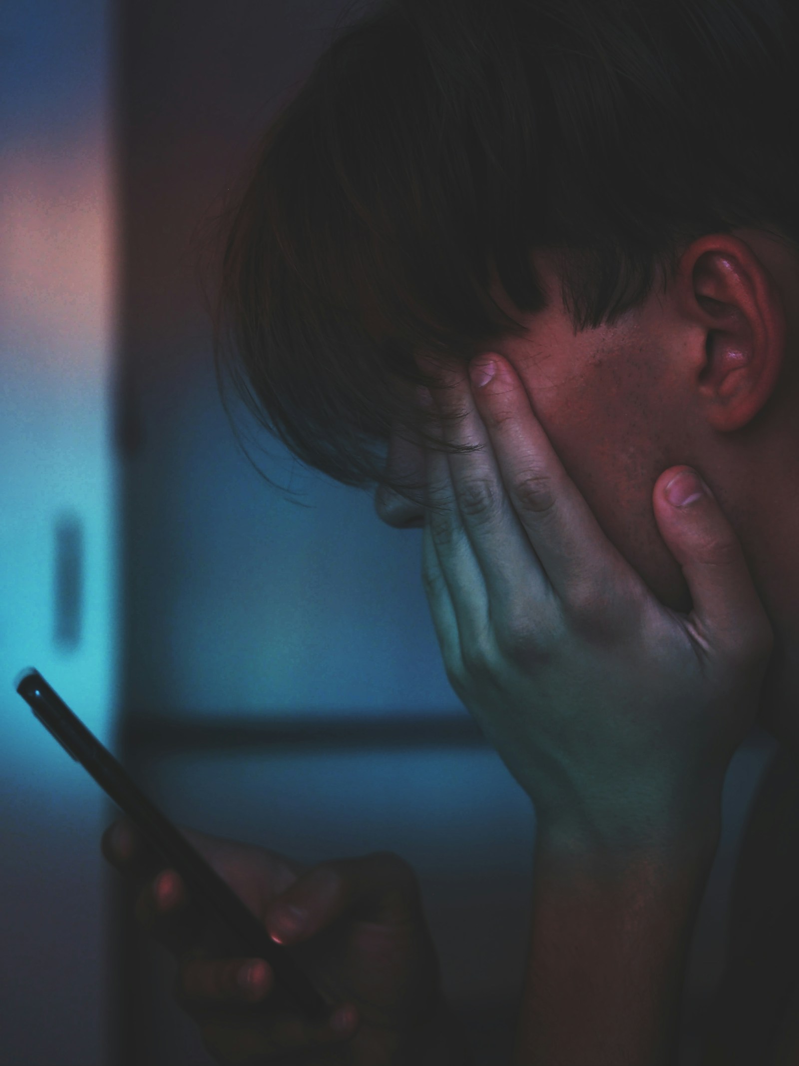 A man in dim light holding his phone with one hand pressed to his face in frustration