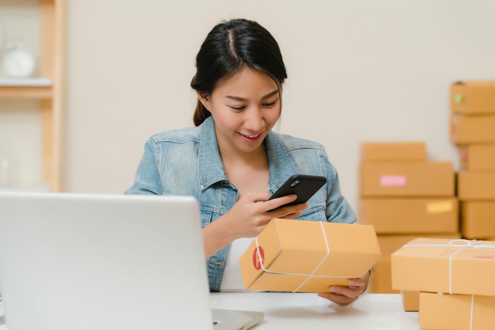 Small online-store owner checking an order on her phone while holding a parcel, with packed boxes behind her