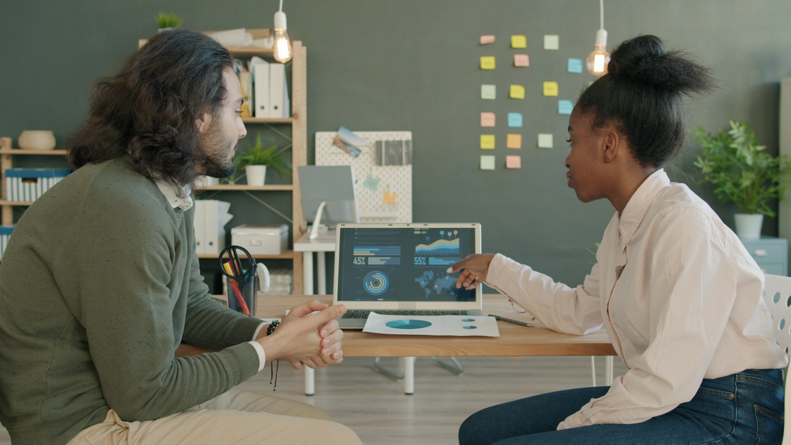 Two colleagues sit at a desk while one points at charts on a laptop screen during a discussion