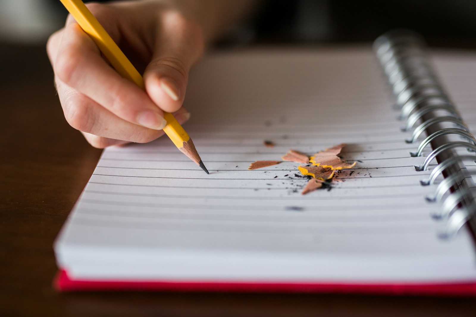 A hand holding a yellow pencil writes on a lined spiral notebook next to fresh pencil shavings
