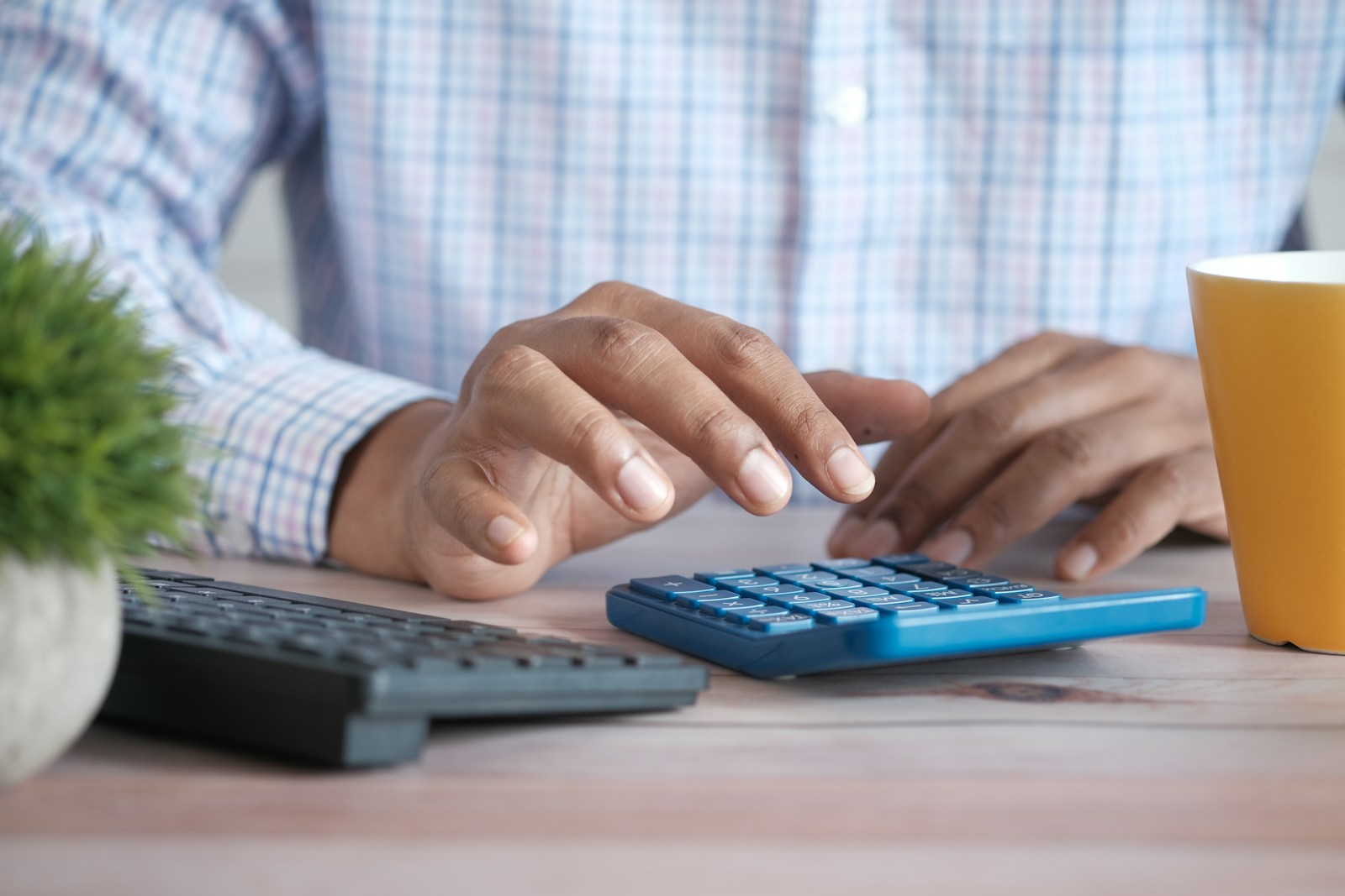 Close-up of a person in a checked shirt pressing keys on a blue calculator beside a keyboard and a yellow mug