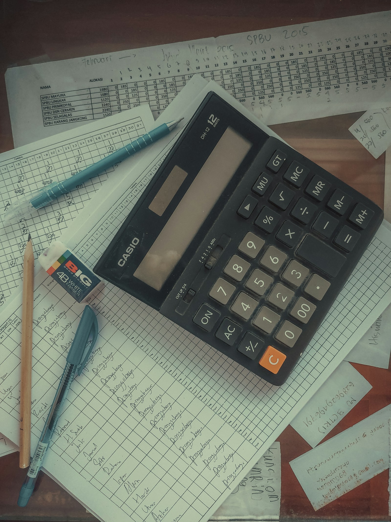 A black desk calculator resting on handwritten ledger sheets and printed spreadsheets, with a pen and pencil beside it