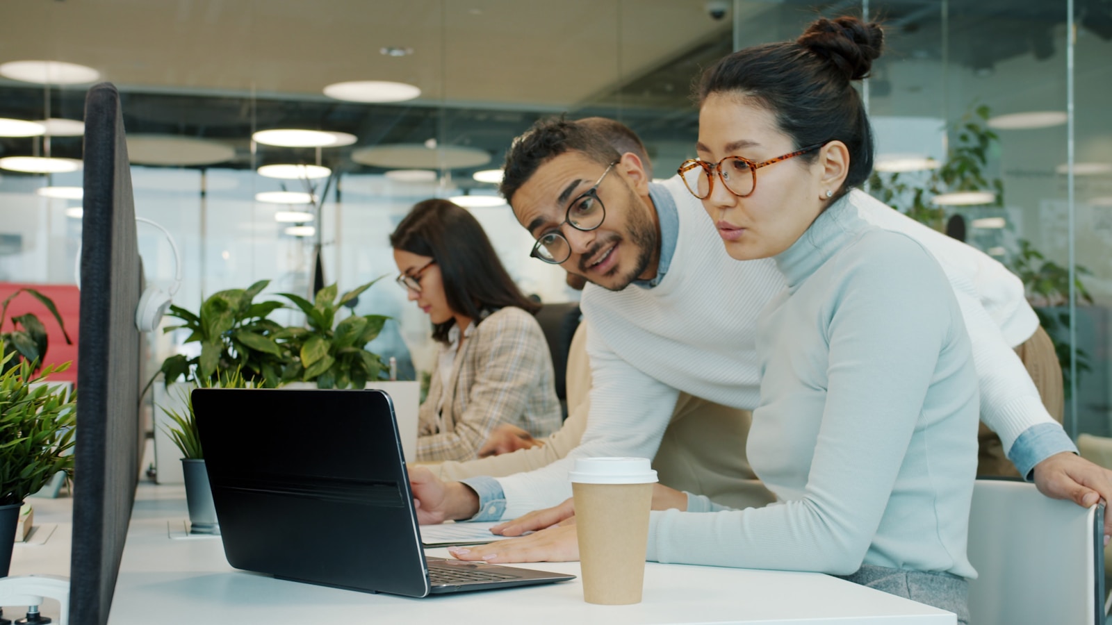 Two colleagues leaning over a laptop in an open-plan office, discussing what is on the screen