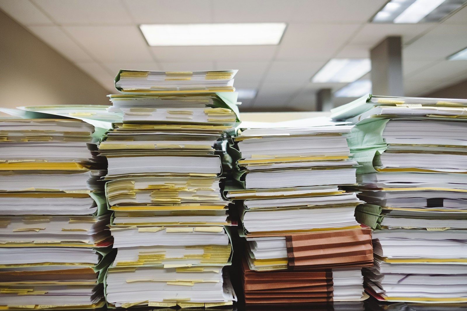 Stacks of paper folders with yellow tabs piled high on an office desk under fluorescent lights