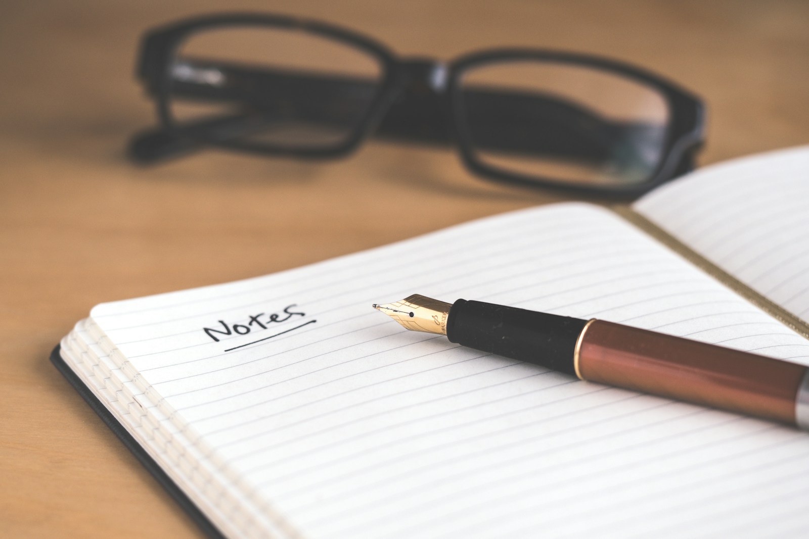 An open lined notebook with the word Notes handwritten at the top, a fountain pen resting on the page and reading glasses behind it